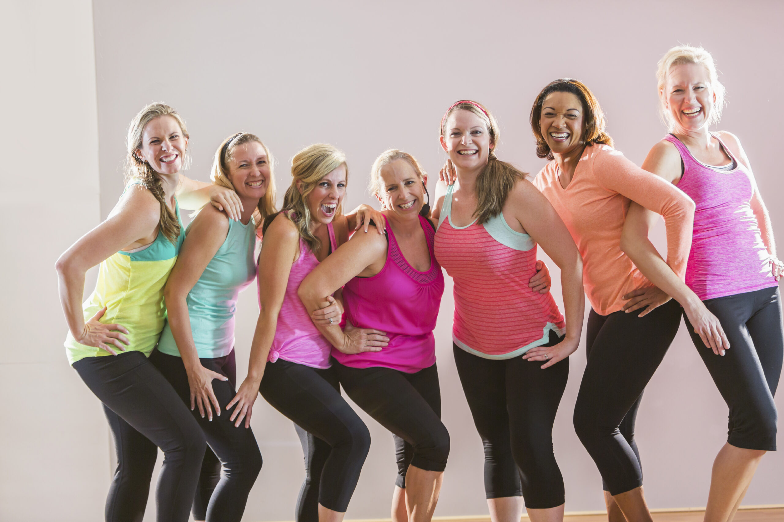 A group of women taking an exercise class, standing in a row posing for the camera.  The instructor, who is wearing a microphone, is standing in the center.